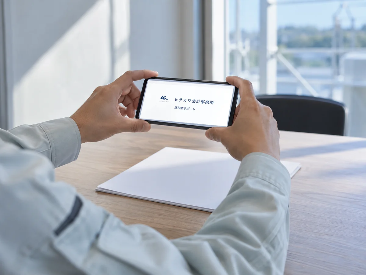 事務所のロゴを表示したスマートフォンを両手で持つ手元